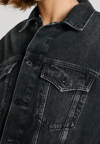 Svart denimjacka med knäppning framtill, två bröstfickor och kontrasterande vit sömnad, med klassisk krage och strukturerad tyg.