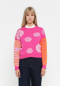 Vaaleanpunainen neulepusero, jossa on kukka- ja eläinprinttejä, kontrastiväriset hiharesorit oranssina ja vaalean violetina, ribbaus kaula-aukossa ja helmassa. Denim-housut.
