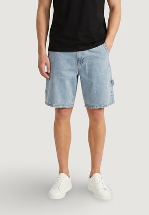 Uomo che indossa pantaloncini di jeans azzurri, maglietta nera, scarpe da ginnastica bianche, in piedi con una mano in tasca davanti a uno sfondo semplice.