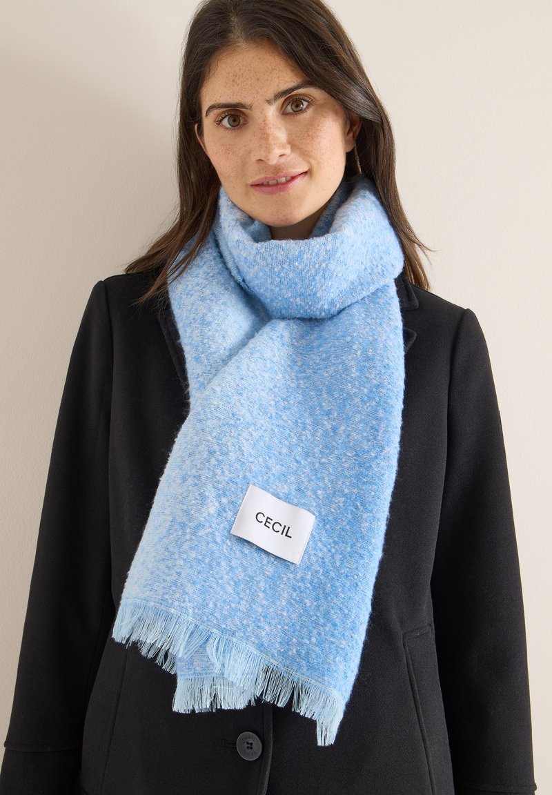 Světle modrý šátek s texturovaným vzorem, třásněmi na okrajích a našitou etiketou s nápisem "CECIL", nošený přes černý kabát.