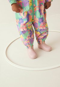 Kolorowy, wzorzysty kombinezon przeciwdeszczowy dla dzieci w kolorze zielonym z różnymi ilustracjami zwierząt. W zestawie różowe, wodoodporne kalosze. Okrągły biały obrys.