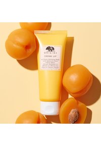 Gele tube van Origins hydraterend masker met een witte dop en label. Er omheen liggen hele abrikozen met zichtbare texturen en pitten.