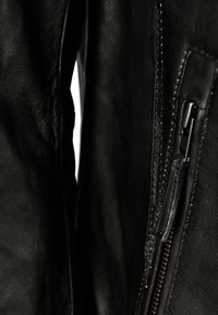 Detail einer schwarzen Lederjacke mit glatter Textur, exponiertem Reißverschluss auf der rechten Seite, kontrastierenden Nähten und subtiler Oberfläche des Materials.