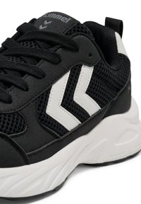 Schwarz-weißer Sportschuh mit Mesh-Gewebe, weißem Chevron-Logo an der Seite, schwarzen Schnürsenkeln und dicker, strukturierter weißer Sohle.