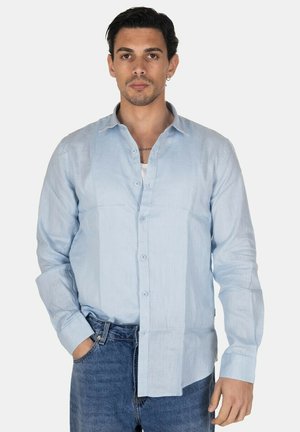 Uomo con capelli scuri indossa una camicia azzurra con bottoni sopra una maglietta bianca, jeans blu, in piedi con una mano in tasca su sfondo semplice.