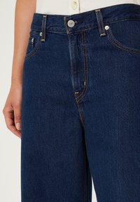 Ciemnoniebieskie dżinsy z prostymi nogawkami, ozdobione kontrastującymi złotymi szwami i metalowym guzikiem zapinanym w pasie.