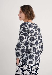 Blumenbluse mit rundem Halsausschnitt, die ein blau-weißes Muster aufweist. Der Stoff wirkt leicht und hat lockere Ärmel.