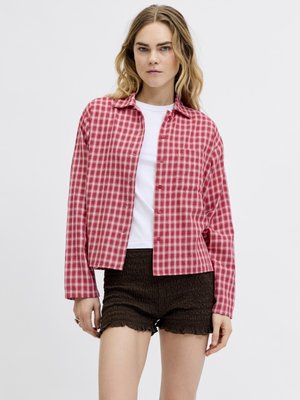 Mujer con camisa de cuadros roja y blanca abotonada sobre una camiseta blanca y shorts negros texturizados, de pie frente a un fondo liso.