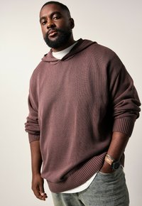 Übergroßer, kastanienbrauner Strickhoodie mit gerippten Bündchen und Saum. Getragen über einem weißen Shirt, kombiniert mit hellblauen Jeans. Hat einen lässigen Schnitt.