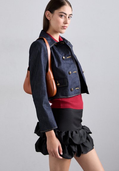 Veste en denim avec quincaillerie dorée, top rouge côtelé et jupe noire à volants. Le mannequin tient un sac en cuir marron, mettant en valeur une tenue superposée.