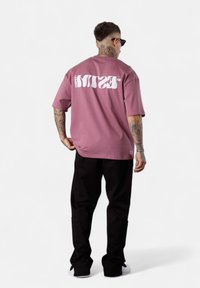 Mann mit Nacken- und Armtattoos, der Sonnenbrillen trägt, ein lockeres mauvefarbener T-Shirt mit weißem Text, schwarze Hosen und weiße Sneakers anhat, steht vor einem weißen Hintergrund.