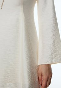 Cremefarbenes Kleid aus strukturiertem Stoff, mit Glockenärmeln und einem dezenten Glanz. Verfügt über vertikale Nähte und eine lockere Passform.