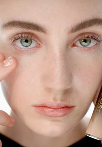 Concealer tube holdt nær et ansikt med lys hud, lyserosa lepper og klare blå-grønne øyne, med fine øyevipper og subtile fregner.