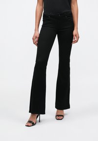 Jeans neri a zampa con vita media, dotati di tasche frontali, una texture liscia e dettagli di cuciture sottili, abbinati a sandali neri con tacco alto.