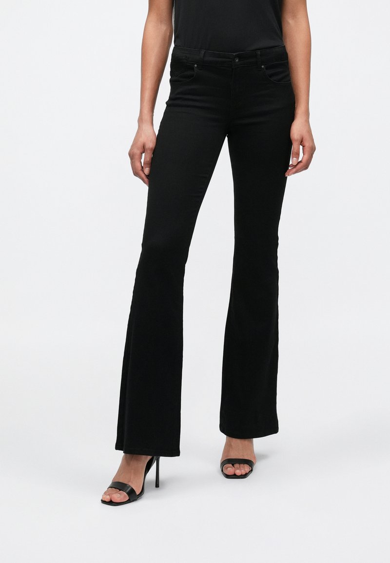 Jeans neri a zampa con vita media, dotati di tasche frontali, una texture liscia e dettagli di cuciture sottili, abbinati a sandali neri con tacco alto.