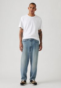 Witte katoenen T-shirt, losse pasvorm. Lichtblauwe denim jeans, rechte pijp, vervaagde afwerking. Schoeisel: zwart en tan sneakers met witte veters.