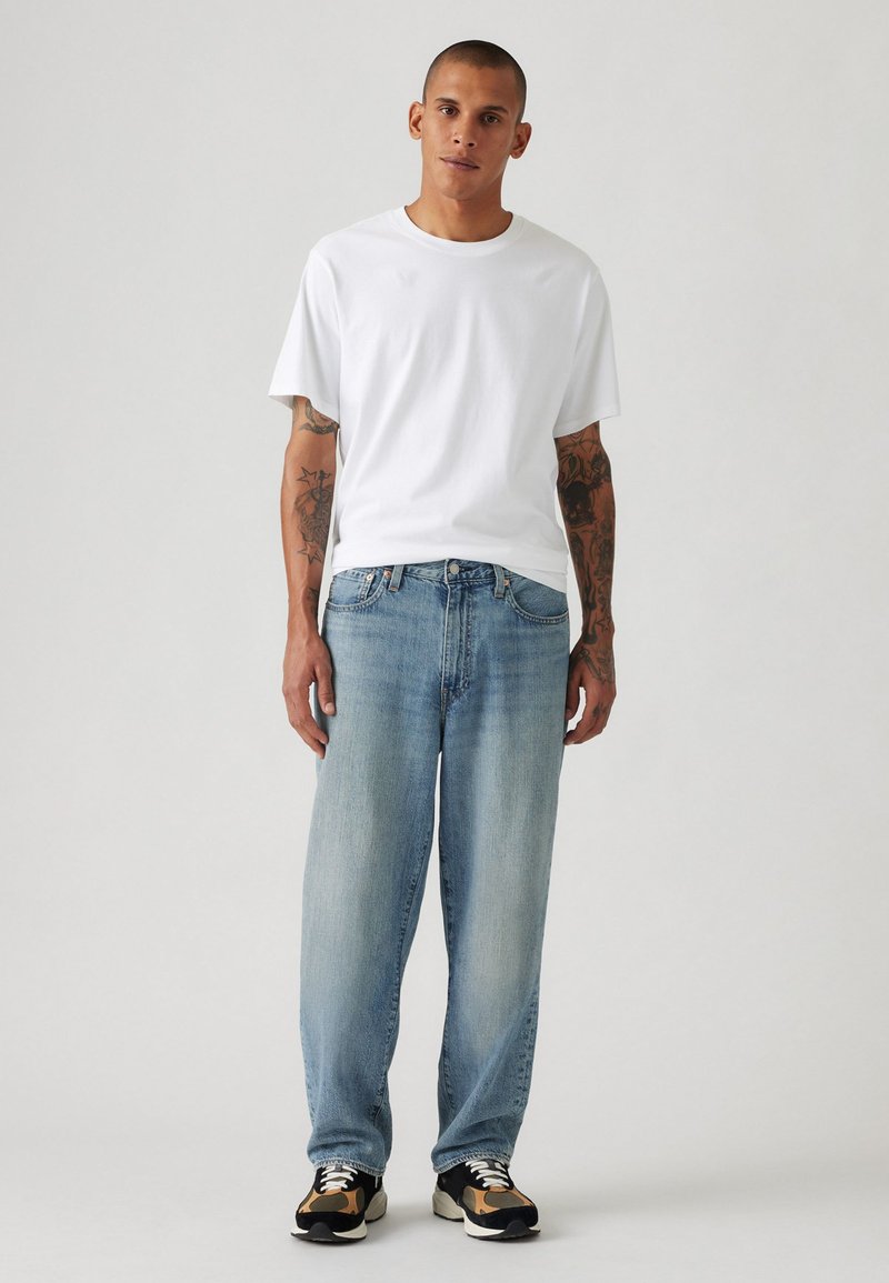 Witte katoenen T-shirt, losse pasvorm. Lichtblauwe denim jeans, rechte pijp, vervaagde afwerking. Schoeisel: zwart en tan sneakers met witte veters.