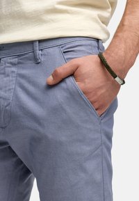 Hellblaue Baumwollhose mit strukturiertem Finish, ausgestattet mit Seitentaschen und einem schmalen Schnitt, ergänzt durch ein grünes geflochtenes Armband.