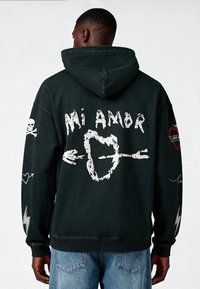 Felpa nera con cappuccio con grafica bianca che riporta il testo "Mi Amor", cuori e design di teschi. Vestibilità rilassata con tasca a marsupio e polsini a coste.
