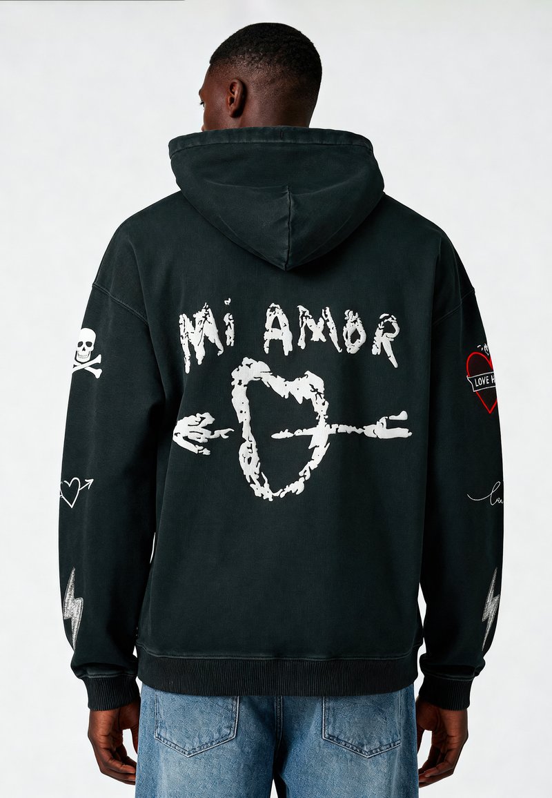 Felpa nera con cappuccio con grafica bianca che riporta il testo "Mi Amor", cuori e design di teschi. Vestibilità rilassata con tasca a marsupio e polsini a coste.