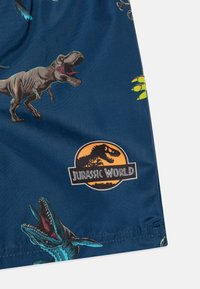 Costumi da bagno blu con stampe di dinosauri, tra cui un T-Rex e un rettile marino, con un logo di "Jurassic World" in arancione e nero.