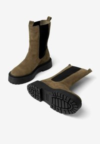 Botas de tobillo de ante marrón con paneles laterales elásticos negros, suelas de goma planas y textura acanalada para mayor tracción. Diseño minimalista.