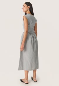 Ärmelloses graues Kleid mit einem eng anliegenden, gerippten Oberteil und einem gerafften Taillenbereich, das unterhalb des Knies endet. Glatter Stoff und schlichtes Design. Schwarze flache Schuhe.