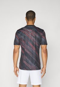 T-shirt de sport à manches courtes en bleu marine avec un motif abstrait rouge et noir, col rond et texture lisse. Des shorts blancs visibles en bas.