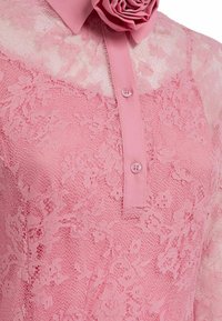 Blusa di pizzo rosa con un'applicazione floreale al colletto, chiusura con bottoni e tessuto testurizzato con motivi floreali.