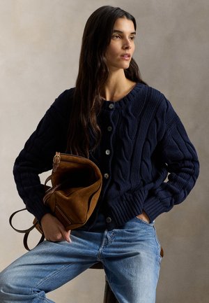 Námornícka pletená kardigán s vrkočovým vzorom a texturovaným stehom, s čiernymi gombíkmi. Neformálne držaná hnedá semišová kabelka.