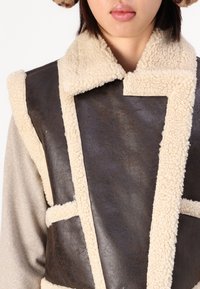 Brun læderveste med cremefarvet shearling-krave og kant, der har et struktureret design og åben front, hvilket skaber en lagdelt teksturkontrast.