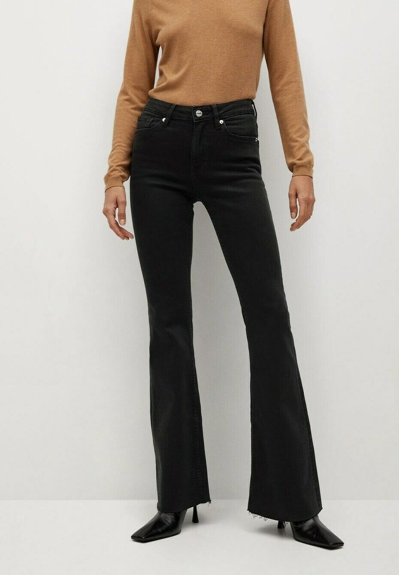 Personne portant un jean flare noir taille haute, un pull à manches longues camel, et des bottes noires à talons pointues sur fond uni.