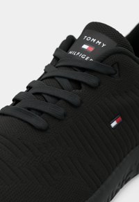 Svart sportskor med en strukturerad ovandel, platta snörningar och en liten logotyp i röd, vit och blå på sidan och tungan.