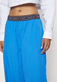 Blå vida byxor i lätt tyg, med ett elastiskt svart midjeband som har "TOMMY JEANS"-logotypen samt röda och vita detaljer.