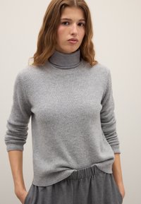 Grey turtleneck sweater made of soft knit fabric. Features ribbed cuffs and hem, with a relaxed fit and subtle texture. Worn with gray pants.
