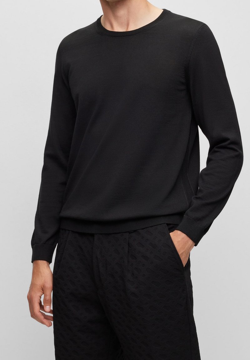 Homme portant un pull noir à manches longues avec col rond et un pantalon noir texturé, une main dans la poche, sur un fond gris clair uni.