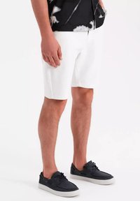 Pantalones cortos de algodón blancos con corte recto, bolsillos laterales y una textura suave, combinados con zapatos casuales de un oscuro azul marino.