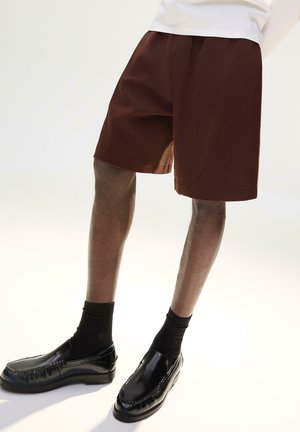 Férfi sötétbarna térdnadrágban, fekete zokniban és fekete bőr mokaszinban, egyszínű világos háttér előtt állva.