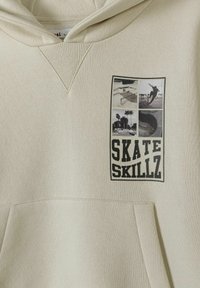 Beige hoodie med känguruficka, med ett grafiskt tryck av skateboardscener och texten "SKATE SKILLZ" i fet svarta bokstäver.
