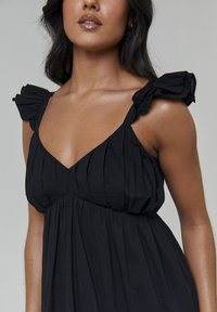 Czarna plisowana sukienka z dekoltem w kształcie litery V i marszczonymi ramiączkami. Miękka faktura i lejąca się sylwetka. Brak widocznych wzorów i ozdób.