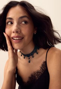 Young woman with short dark hair wearing a black lace top and ornate black choker, smiling and touching her face while looking sideways.