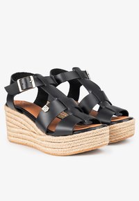 Sandalias de cuña de piel negra con correas entrecruzadas y plataforma de cuerda de yute. Presentan un cierre de hebilla y una plantilla en tono marrón.