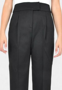 Pantaloni neri su misura con pinces, una cintura larga e una texture liscia, mostrati dalla vita in giù, con le mani sui fianchi.