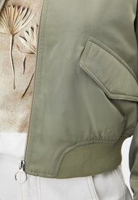 Veste bomber verte avec une fermeture éclair et une poche à rabat portée par-dessus une chemise beige ornée d'un motif de plante séchée, associée à un pantalon blanc.