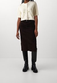 Cremfarbener, bestickter Cardigan mit kurzen Ärmeln und Knöpfen in der Front. Brauner Cord-Midirock mit einer mittigen Naht. Schwarze kniehohe Stiefel.