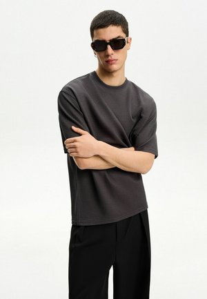 Szürke, rövid ujjú, textúrázott póló kerek nyakkal, fekete, szűk szabású nadrággal és sötét napszemüveggel párosítva. A modell karjára tett kezekkel áll.