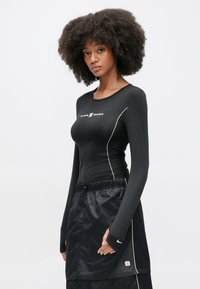 Μαύρο εφαρμοστό τοπ με μακριά μανίκια, λευκό λογότυπο και πλάγια διακριτικά στοιχεία, συνδυασμένο με μαύρη φούστα με ελαστική μέση και λευκή διακόσ�μηση.