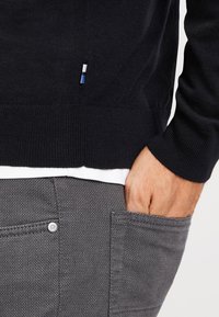 Marineblauwe gebreide trui met een subtiele textuur, voorzien van een klein blauw-wit-blauw accenten label. Grijze broek met patroon en een knoopdetail.