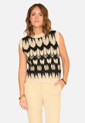 Pull sans manches en tricot beige et noir avec un motif géométrique à finition texturée. Associé à un pantalon en velours côtelé beige clair.
