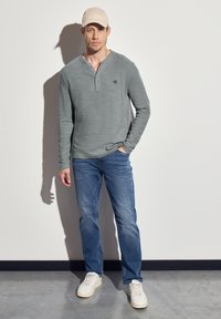 Harmaa pitkähihainen henley-paita, jossa on teksturoitu pinta, ja jossa on rintalogot. Yhdistetty sinisiin farkkuihin ja valkoisiin lenkkarikenkiin. Päällä kerma-tönkkä.